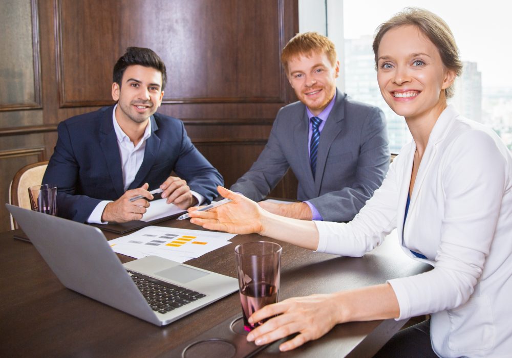 Group of three business people having meeting in conference room, cheerful businesswoman pointing to laptop screen and discussing business plan with male colleagues